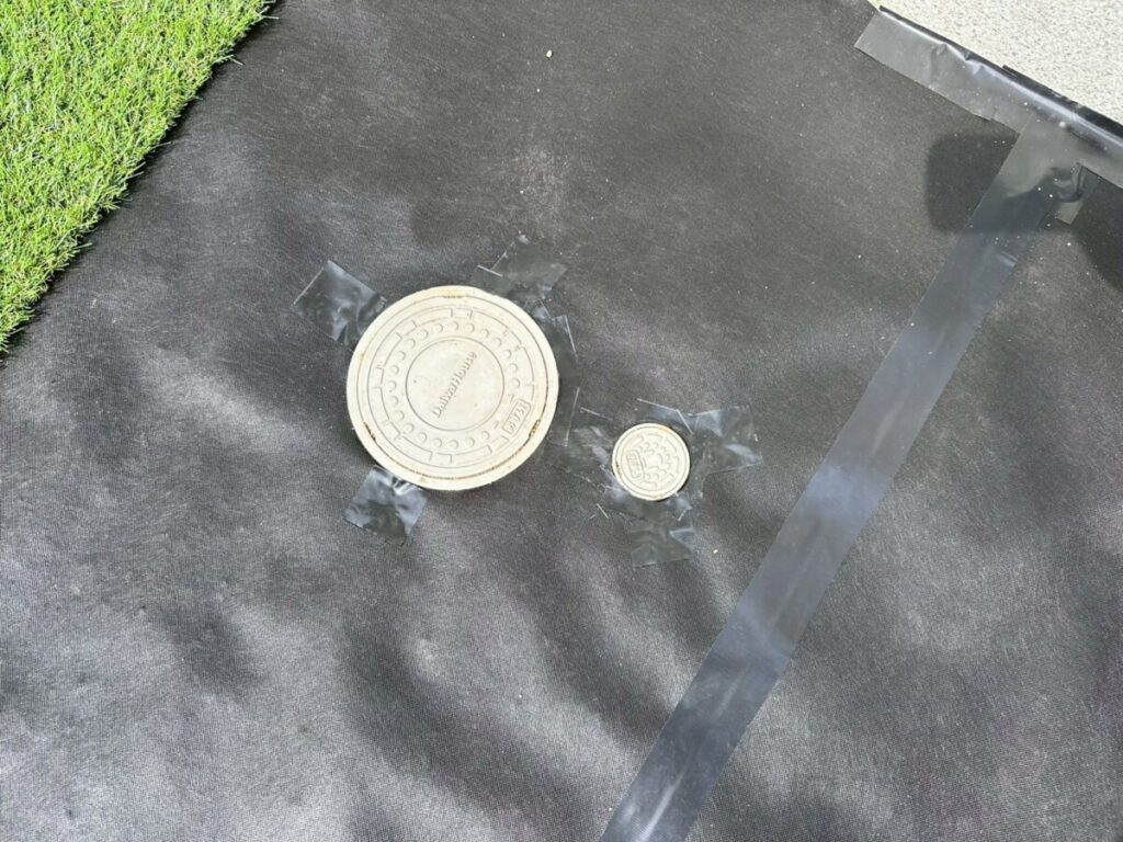 円形の雨水マスの形に合わせて防草シートをハサミで丸くくり抜き、そのキワを黒い専用の粘着テープで隙間なく密閉しているクローズアップ写真