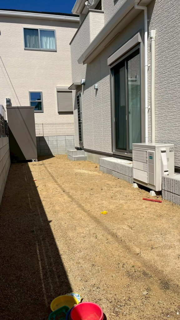 施工前の庭を家側から見たところ。掃き出し窓と室外機の先に土の地面が続き、奥に物置が見える