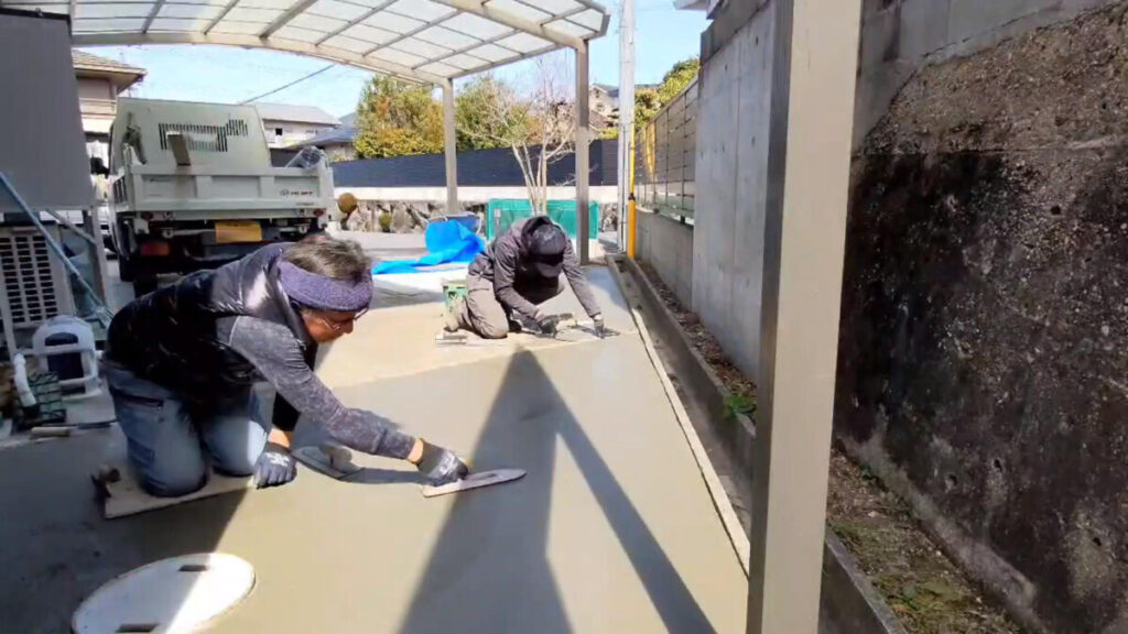 流し込まれたグレーの生コンクリートの表面を、職人が金コテを滑らせてツルツルに仕上げている打設シーン