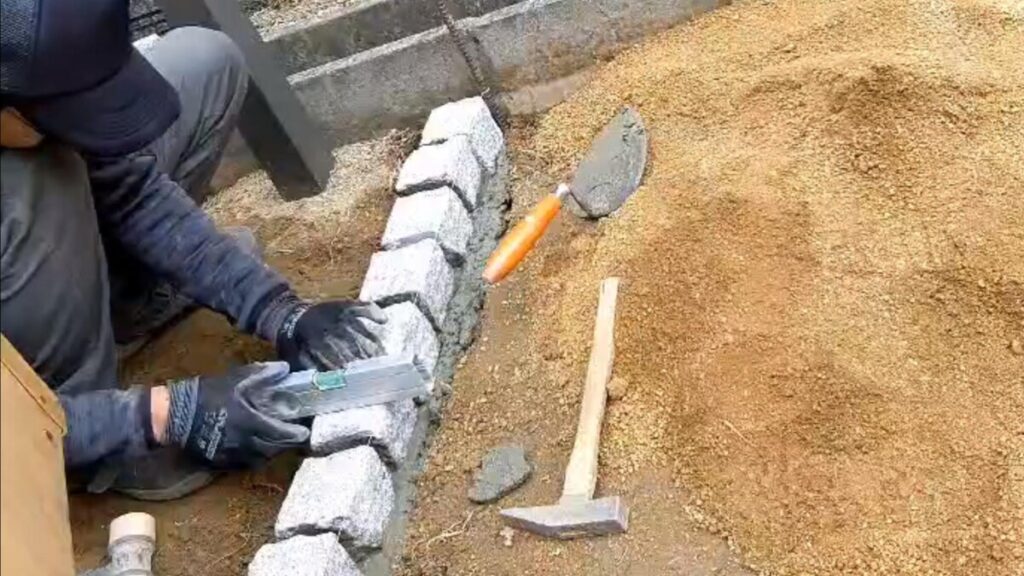 張られた水糸に合わせて、職人がピンコロ石をモルタルで等間隔に美しく据え付けている手元のクローズアップ写真