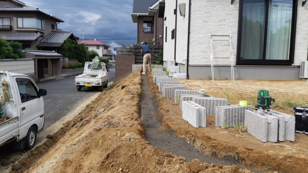 法面の上の縁に、目隠しフェンスの基礎の溝を掘っているところ。手前にブロックの材料