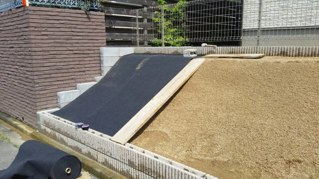 法面に防草シートを敷いているところ。板を当てて押さえながら張り、右側はまだ真砂土のまま
