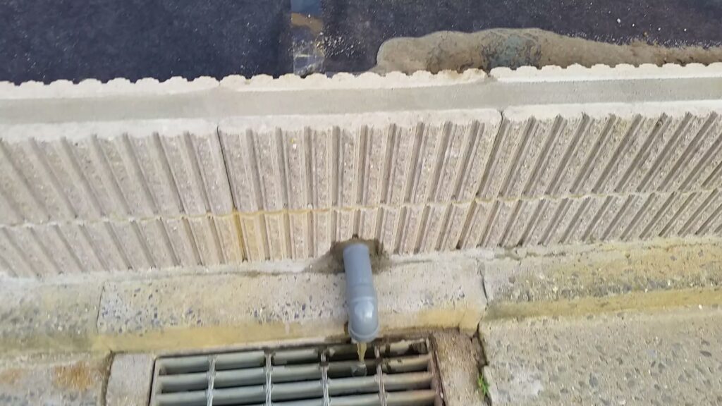 土留めブロックから出た排水管の先から、たまっていた水が道路側の側溝へ流れ落ちているところ
